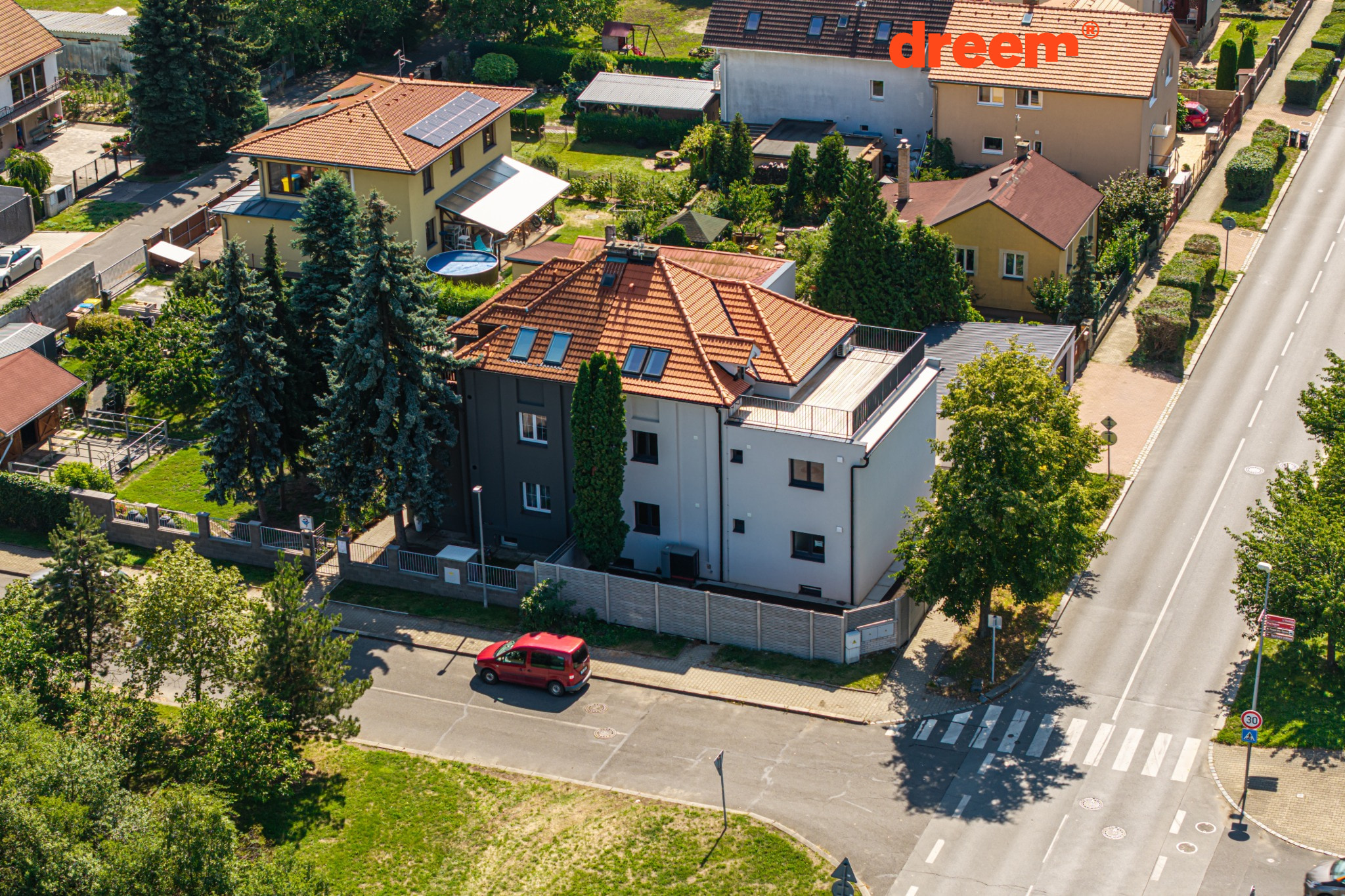 Letecký snímek bytového domu s červenou střechou a okolní zástavbou u silnice