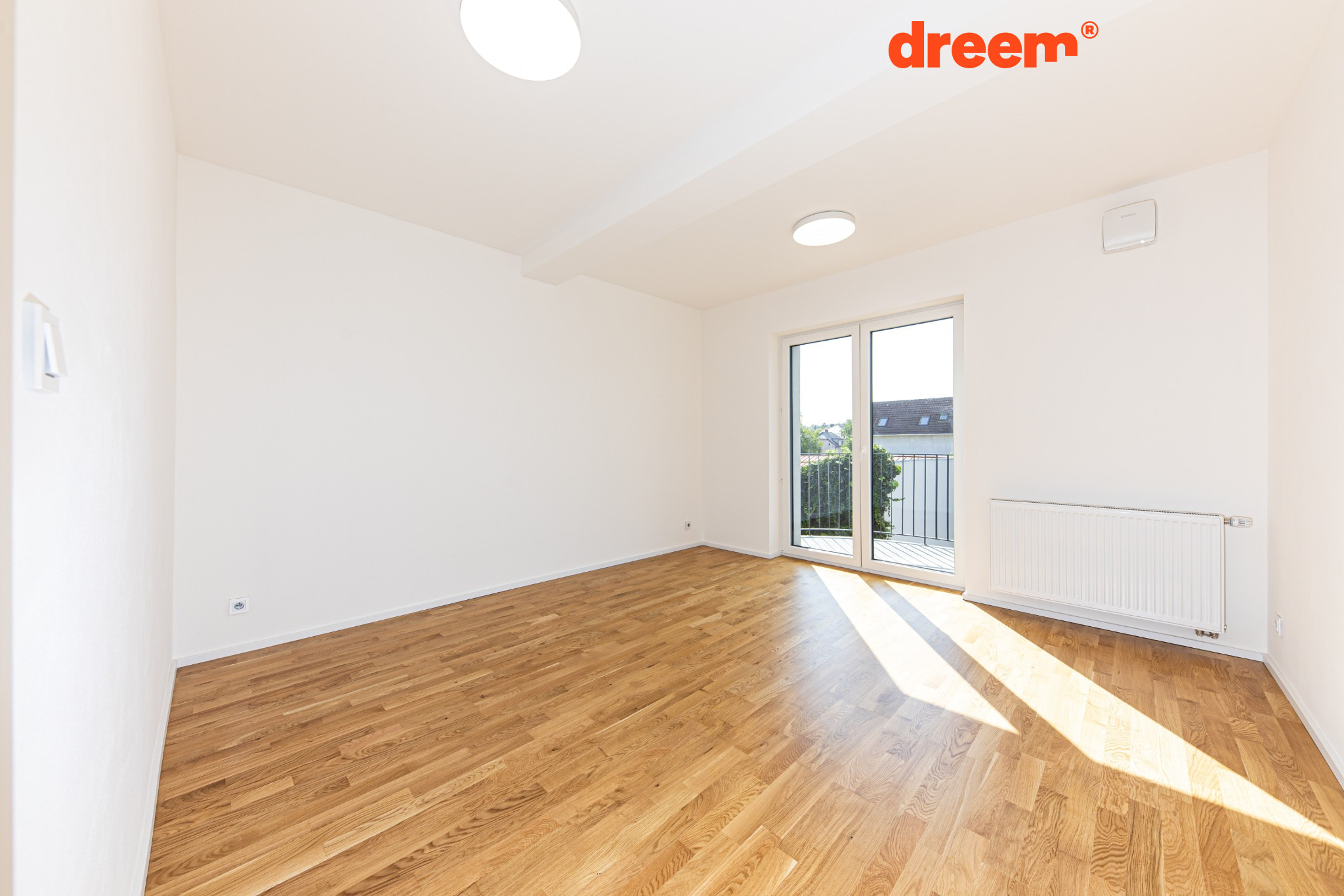 Prázdný pokoj s dřevěnou podlahou, bílými stěnami a prosklenými dveřmi na balkon zalitý sluncem