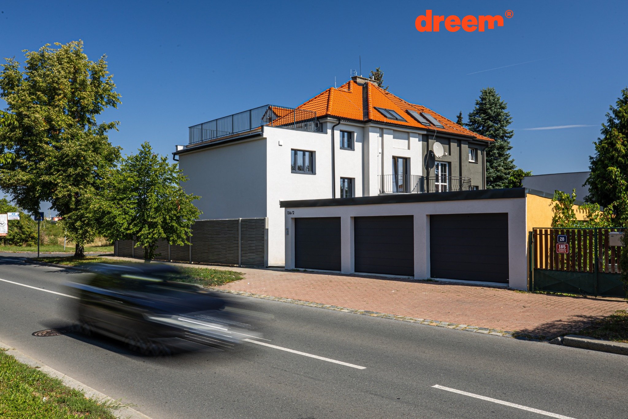 Vícepodlažní rodinný dům se světlou fasádou, oranžovou střechou, třemi garážovými vraty v popředí a projíždějícím rozmazaným autem na silnici.