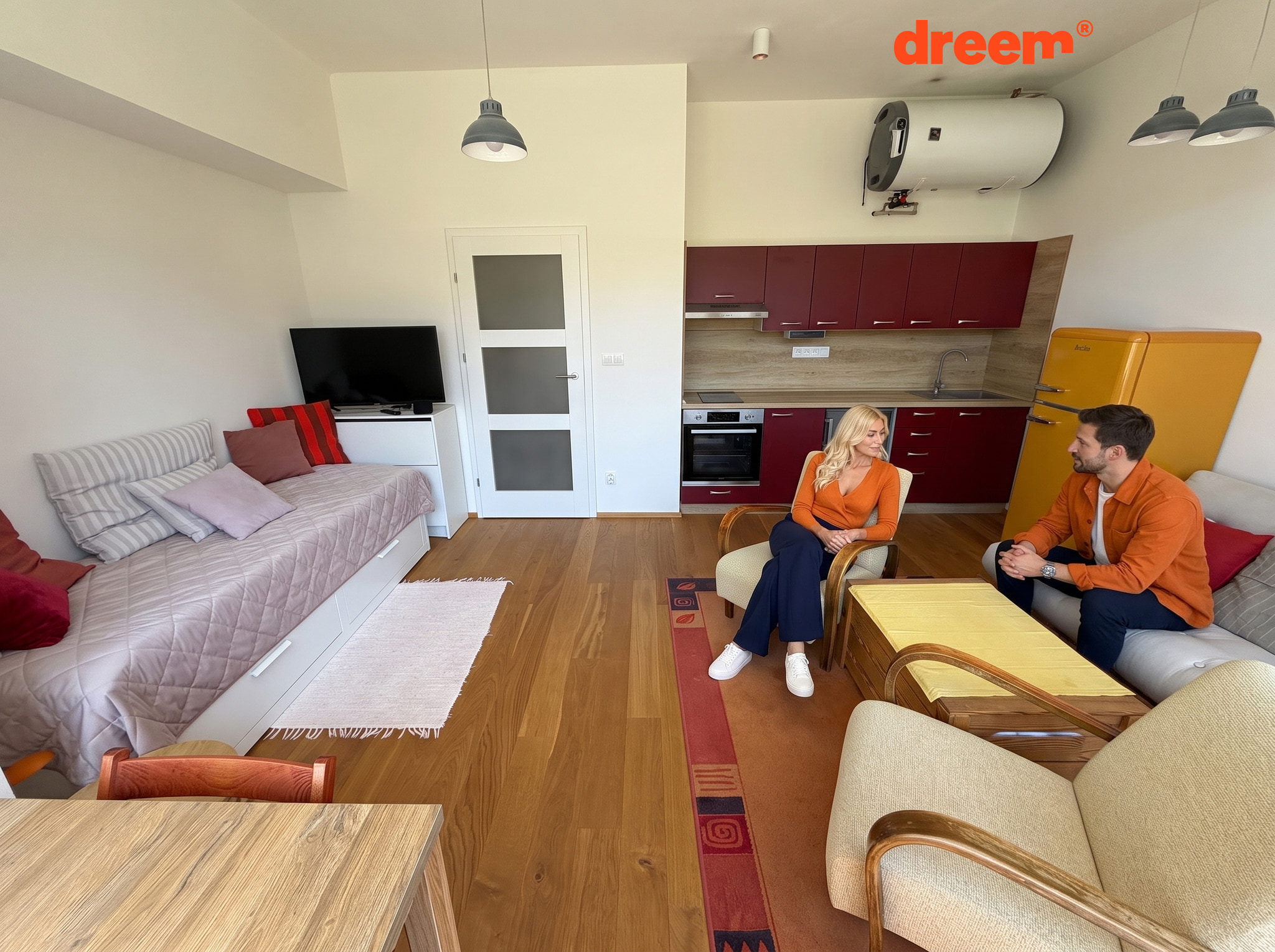 Světlý obývací pokoj propojený s tmavě červenou kuchyňskou linkou, žlutou lednicí, pohovkami a křesly na dřevěné podlaze