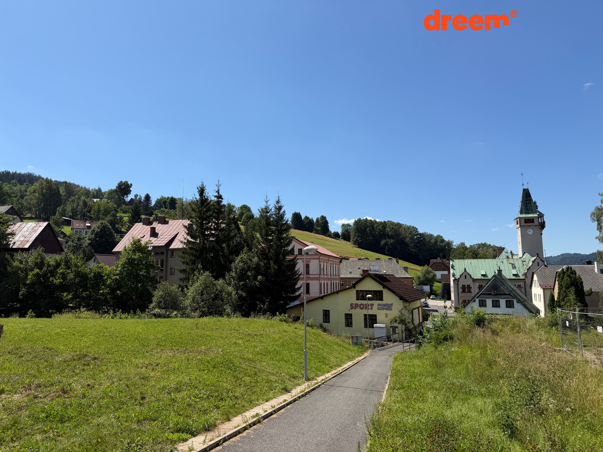 Cesta vedoucí travnatým svahem směrem k budovám v obci s věží v pozadí za jasného dne