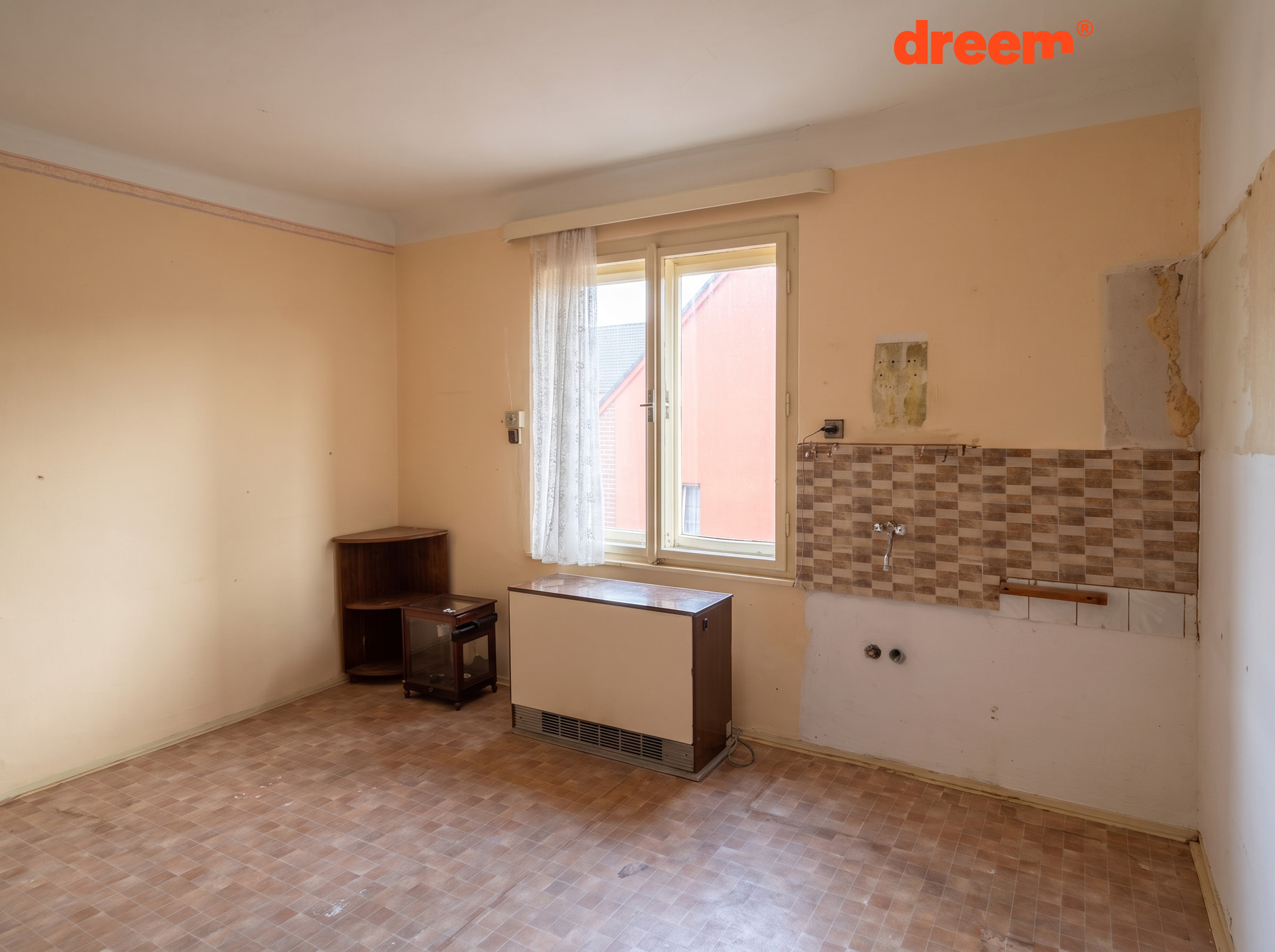 Prázdná místnost s linoleem, oknem a obloženým prostorem pro kuchyňskou linku s vývody vody