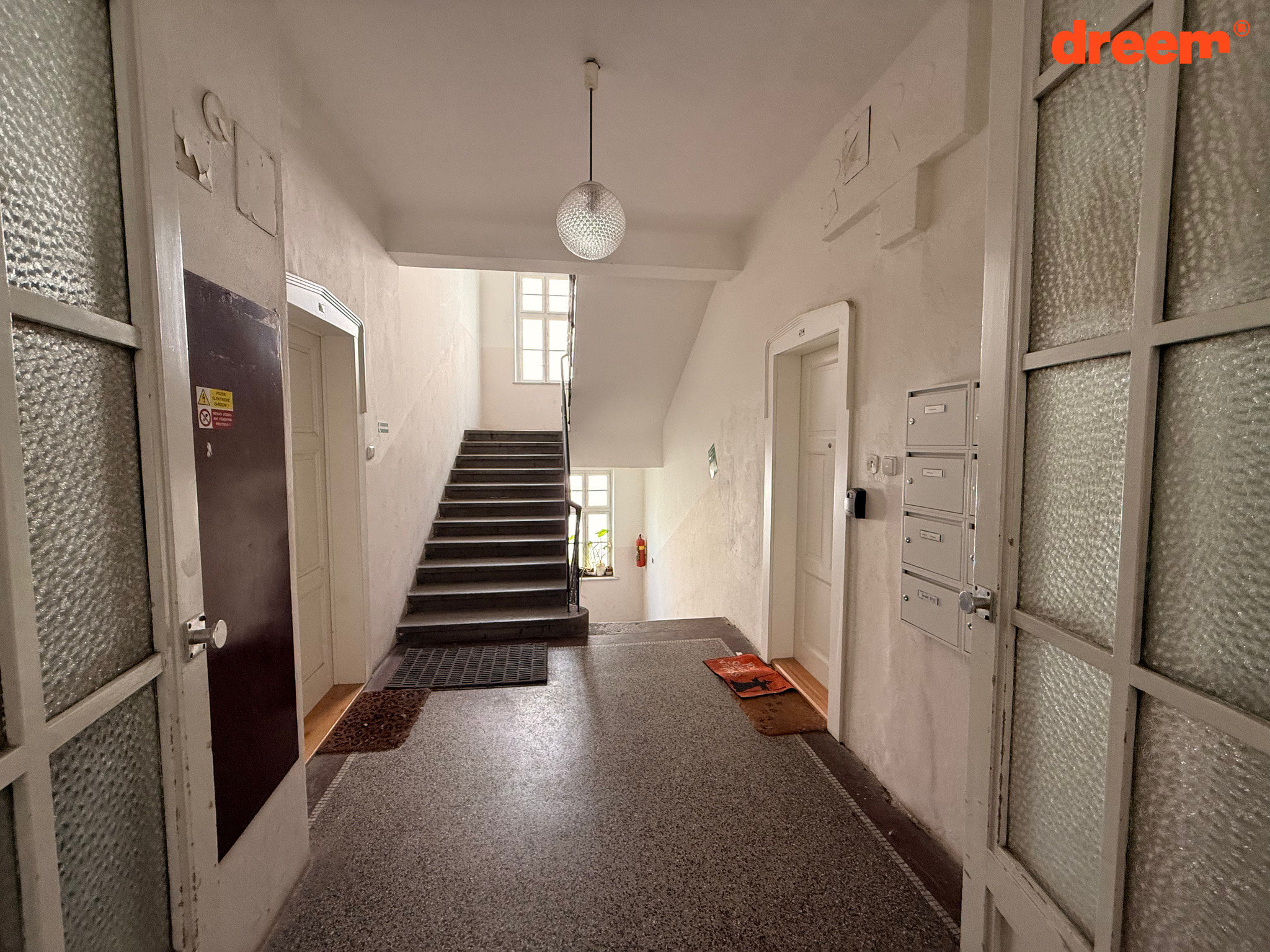 Vstupní chodba bytového domu s teracovou podlahou, schodištěm do patra, poštovními schránkami a vstupními dveřmi do bytů.