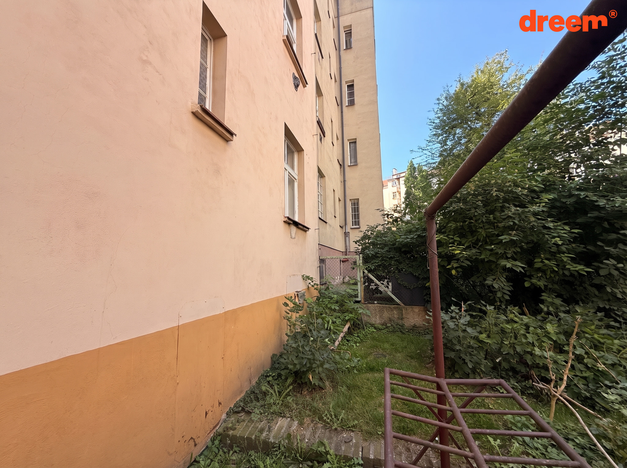 Pohled podél fasády bytového domu do travnatého dvorku se vzrostlou zelení a kovovou konstrukcí.