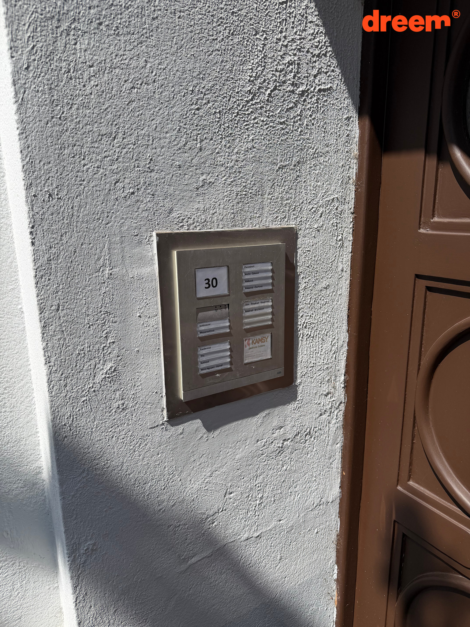 Detailní pohled na kovový panel se zvonky a číslem popisným vedle hnědých vchodových dveří.