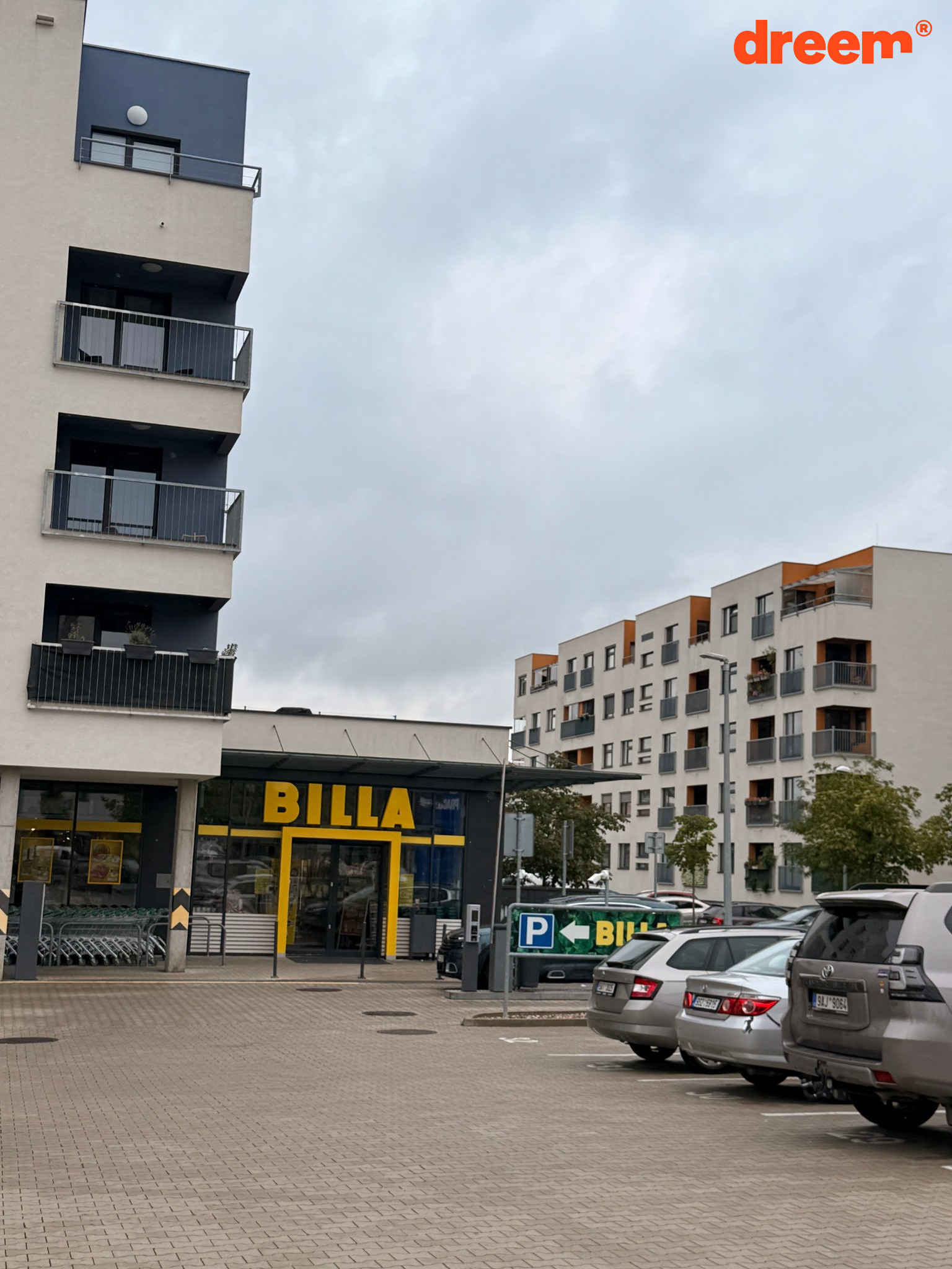 Pohled na parkoviště před prodejnou potravin vedle moderního bytového domu za oblačného dne